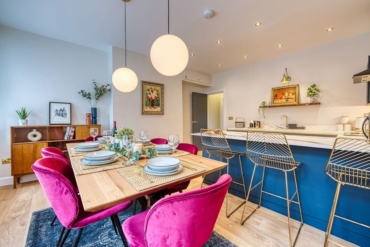 Una cocina moderna con una isla de madera, taburetes de bar y una zona de comedor con mesa de madera y sillas rosas.