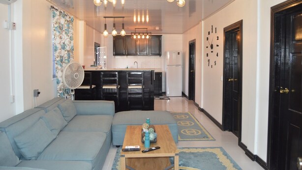 A living room with a sofa, a coffee table, a fan, and a kitchen area with cabinets and a refrigerator. - Apartment Luxe Next to Malls Central Location (Georgetown)