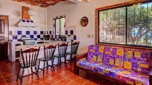 Una cocina con una isla central, sillas de madera, un sofá colorido y un amplio ventanal con vistas a árboles.