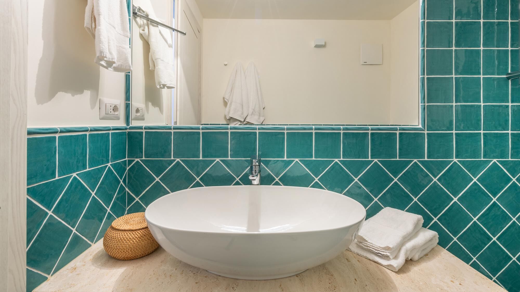 A bathroom with a white sink, teal mosaic tiles, a white towel, and a woven basket on the counter.