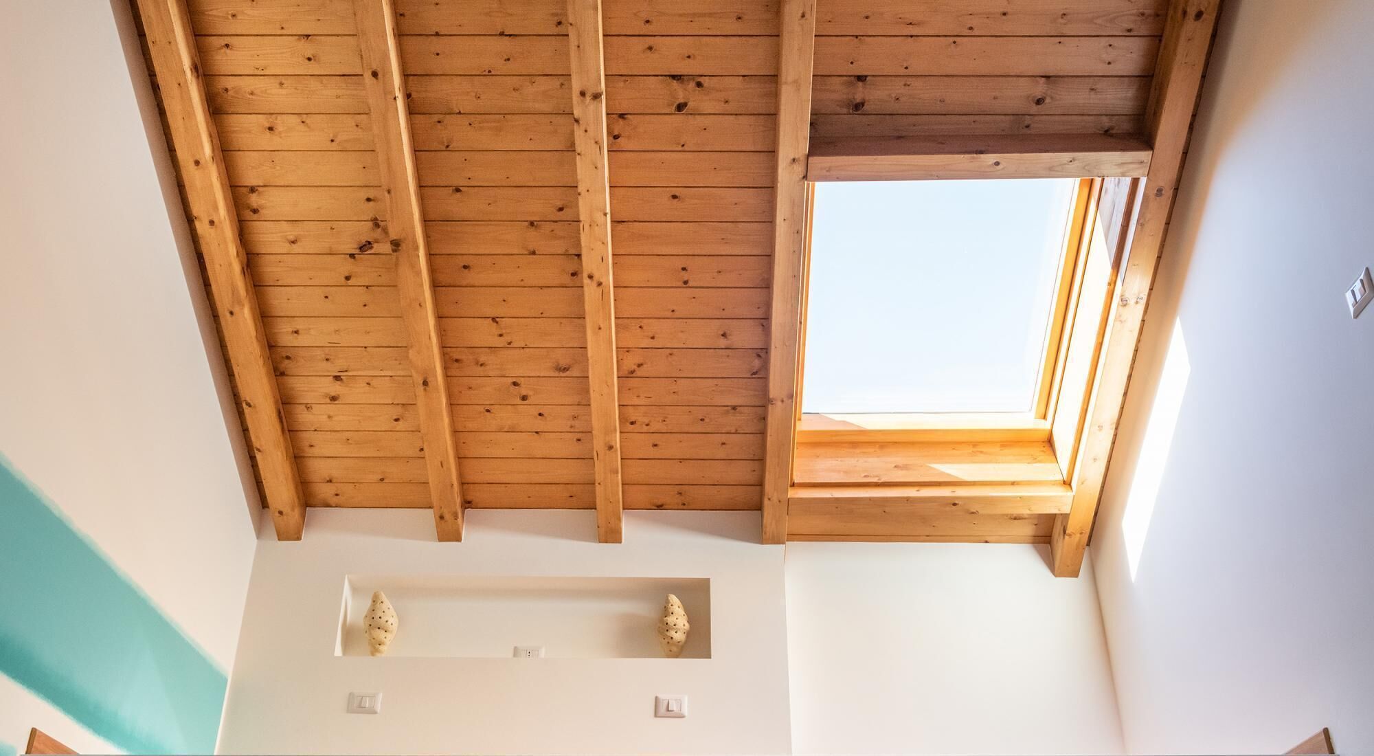 Une pièce avec un plafond en pente, des poutres en bois, un puits de lumière et un mur blanc avec deux interrupteurs.