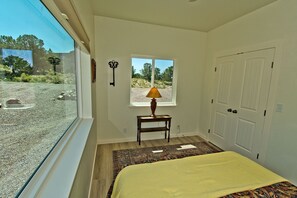 A bedroom with a large window, a bed with a yellow bedspread, a small table with a lamp, and a door. - Challenger Views on Ridgecrest (Saguache County)