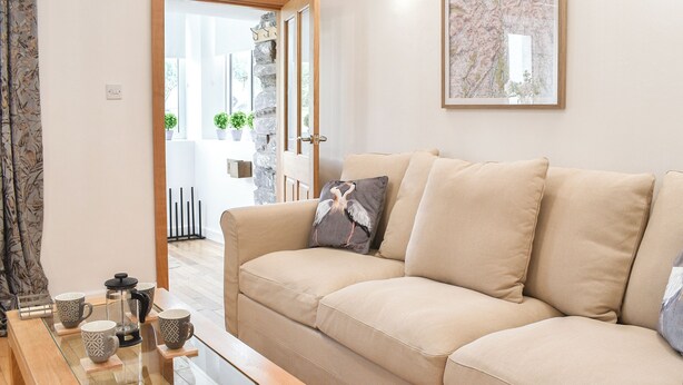 Una sala de estar con un sofá color beige, una mesa de centro de vidrio y una puerta que da a un balcón con plantas.