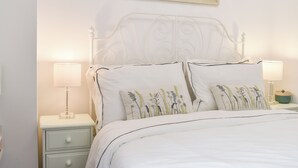Una cama bien hecha con almohadones florales, un cabecero blanco y lámparas de noche sobre una mesita blanca.
