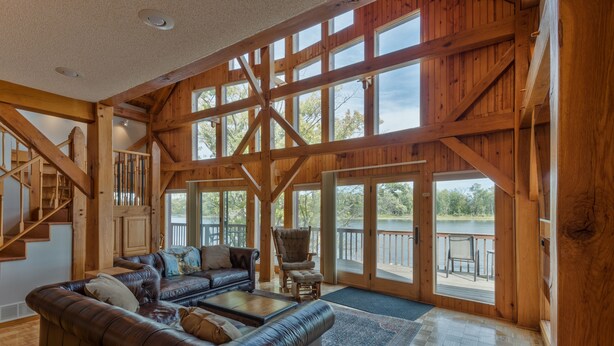 A spacious living room with a large wooden staircase, a leather sofa, and a view of a lake through large windows. - Water Street Lodge! Waterfront 4 Bedroom, 4 Bath, Sleeps 12! (Marquette)