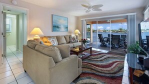 A living room with a sofa, coffee table, and lamps. There is a dining area with chairs and a table. A painting is on the wall, and a ceiling fan is visible. - Casa Laura Unit B - Lake Louise with a private pool (Cape Coral)