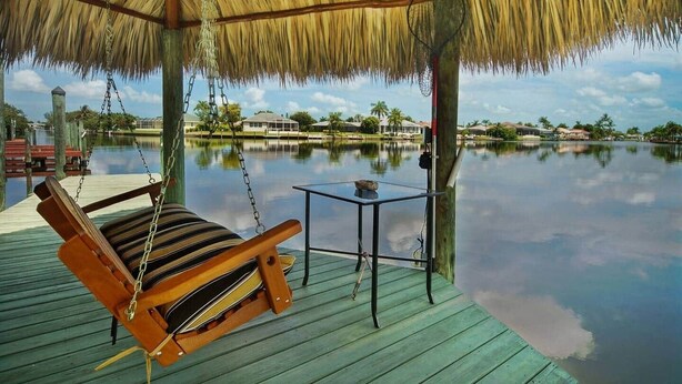 A wooden swing chair under a thatched roof, a small table, and a calm water body with houses in the background. - Casa Laura Unit B - Lake Louise with a private pool (Cape Coral)