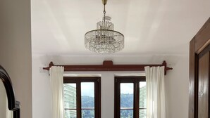 A bedroom with a bed, two windows with curtains, a radiator, a small table with a plant, and a chandelier.