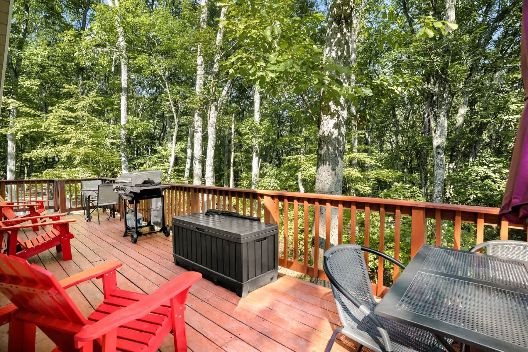Puinen terassi, jossa on punaiset Adirondack-tyyliset tuolit, grilli ja musta säilytyslaatikko.