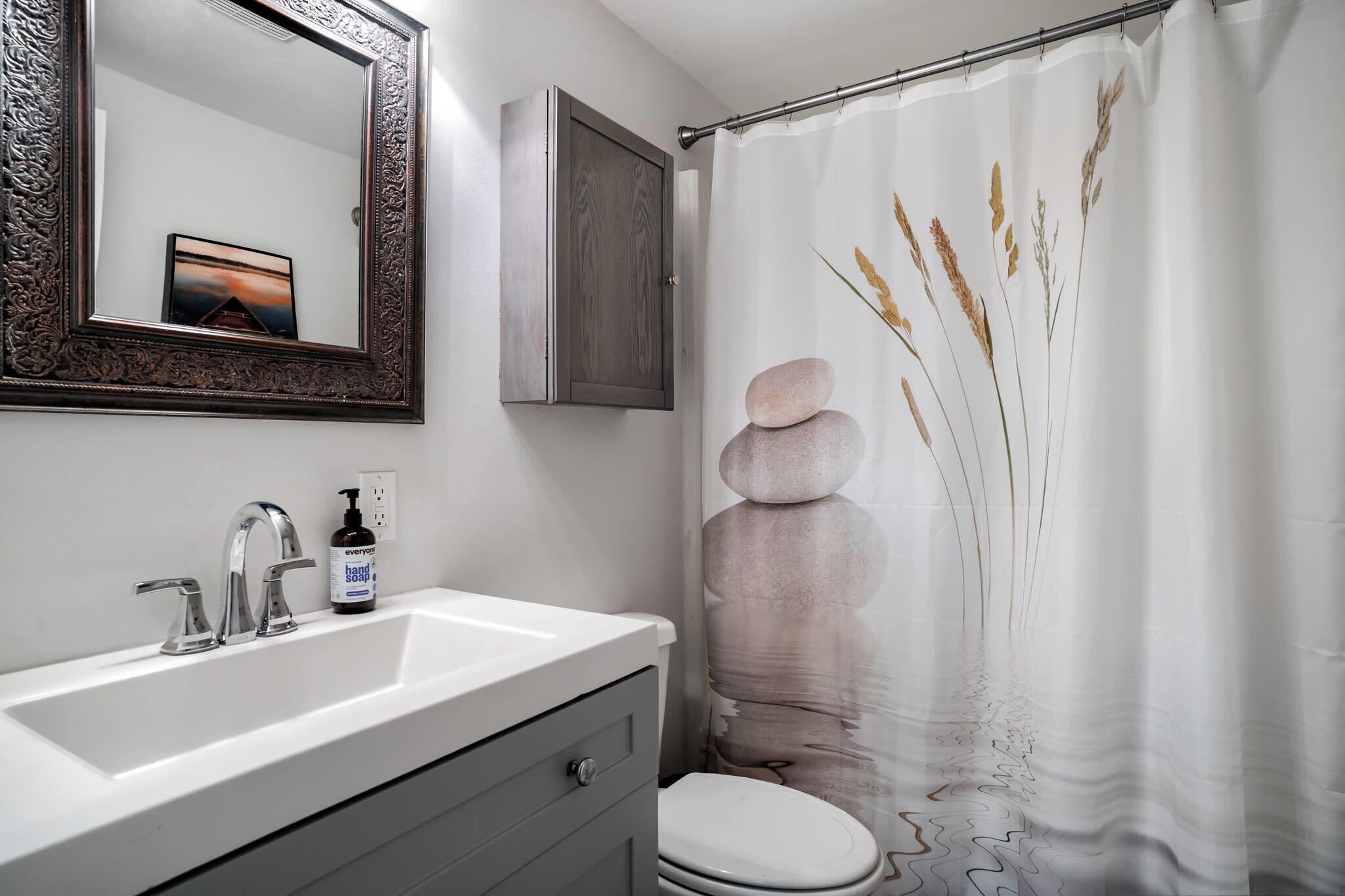 A bathroom with a white sink, a mirror with an ornate frame, a cabinet, a toilet, and a shower curtain with a nature design.