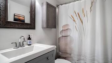 A bathroom with a white sink, a mirror with an ornate frame, a cabinet, a toilet, and a shower curtain with a nature design.