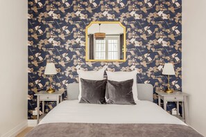 Une chambre à coucher avec un lit, deux tables de chevet, des lampes, un miroir et un papier peint à motifs.