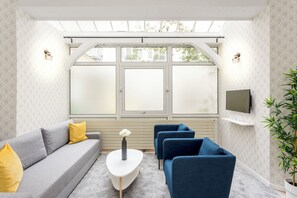 Un salon moderne avec un canapé gris, des fauteuils bleus, une table basse blanche et une grande fenêtre dotée de panneaux de verre dépoli.
