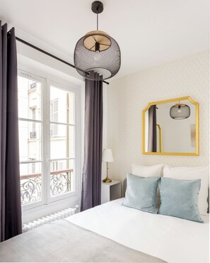 Une chambre d’hôtel avec un lit, une lampe de chevet, un miroir rond avec un cadre doré et une fenêtre avec des rideaux.