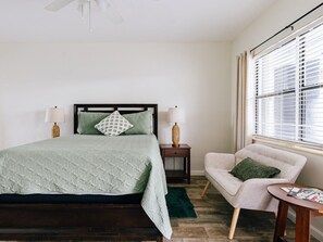 A bedroom with a bed, bedside lamps, a chair, a small table, and a window with blinds.