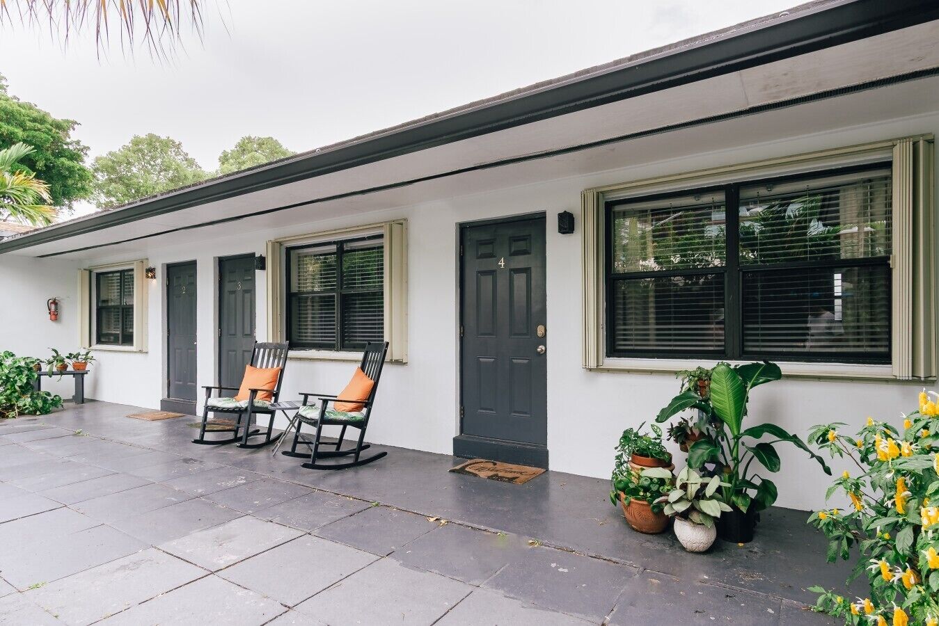 A building with a white exterior, a black door, and windows with blinds. There are two rocking chairs with orange cushions outside.