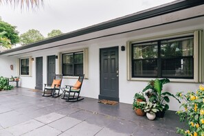 A building with a white exterior, a black door, and windows with blinds. There are two rocking chairs with orange cushions outside.