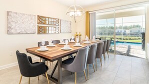 Un comedor con una larga mesa de madera dispuesta para seis personas, rodeada por sillas grises con patas doradas. Hay dos grandes pinturas abstractas en la pared, un candelabro encima y vista a una piscina a través de puertas de vidrio amplias.