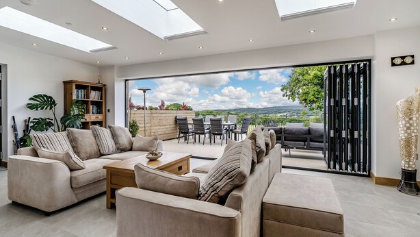 A spacious living room with a large sofa set, a coffee table, and a bookshelf. There is a sliding glass door leading to an outdoor patio area with a table and chairs. - 3 bedroom accommodation in Pontyates (Pontyates)