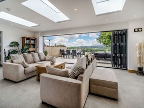 A spacious living room with a large sofa set, a coffee table, and a bookshelf. There is a sliding glass door leading to an outdoor patio area with a table and chairs.