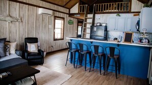 Una habitación acogedora tipo cabaña con techo de madera, una zona de comedor con taburetes azules y una cocina con armarios blancos, además de un pez grande en la pared.