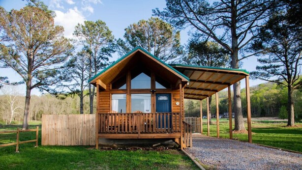 Una cabaña de madera con un porche, rodeada de árboles y un césped.