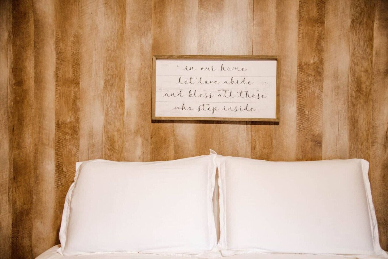 A bed with white bedding against a wooden wall with a framed inspirational quote.