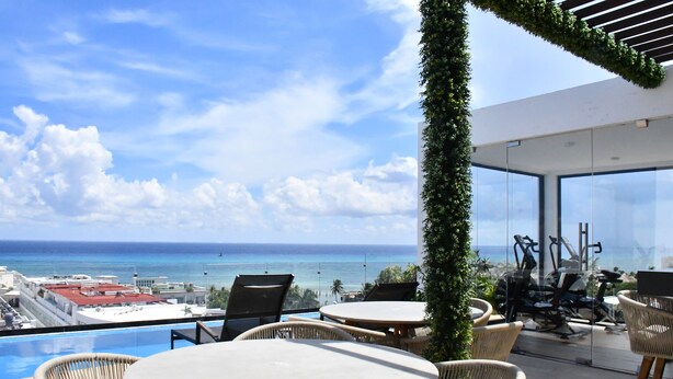 A rooftop pool area with outdoor seating and exercise equipment, overlooking a coastal view. - Luxury Apartment on 5th Av -One Paralia (Playa del Carmen)
