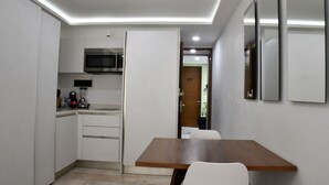 A compact kitchen with white cabinets, a microwave, and a coffee maker. There is a wooden table with chairs in the dining area. - Luxury Apartment on 5th Av -One Paralia (Playa del Carmen)