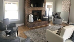 Ein Wohnzimmer mit Kamin aus Backstein, grauen Sofas, einem grauen Teppich und einer Stehlampe.