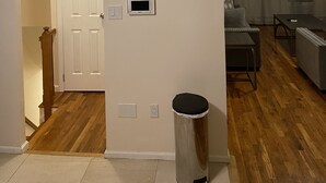 Een woonkamer met houten vloer, een witte deur, een thermostaat, een prullenbak en een eethoek met een tafel en stoelen.