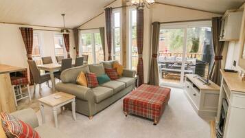 A cozy living room with a sofa, ottoman, and dining table. There is a kitchen area with a stove and cabinets. Large windows provide natural light and a view of the outdoors.