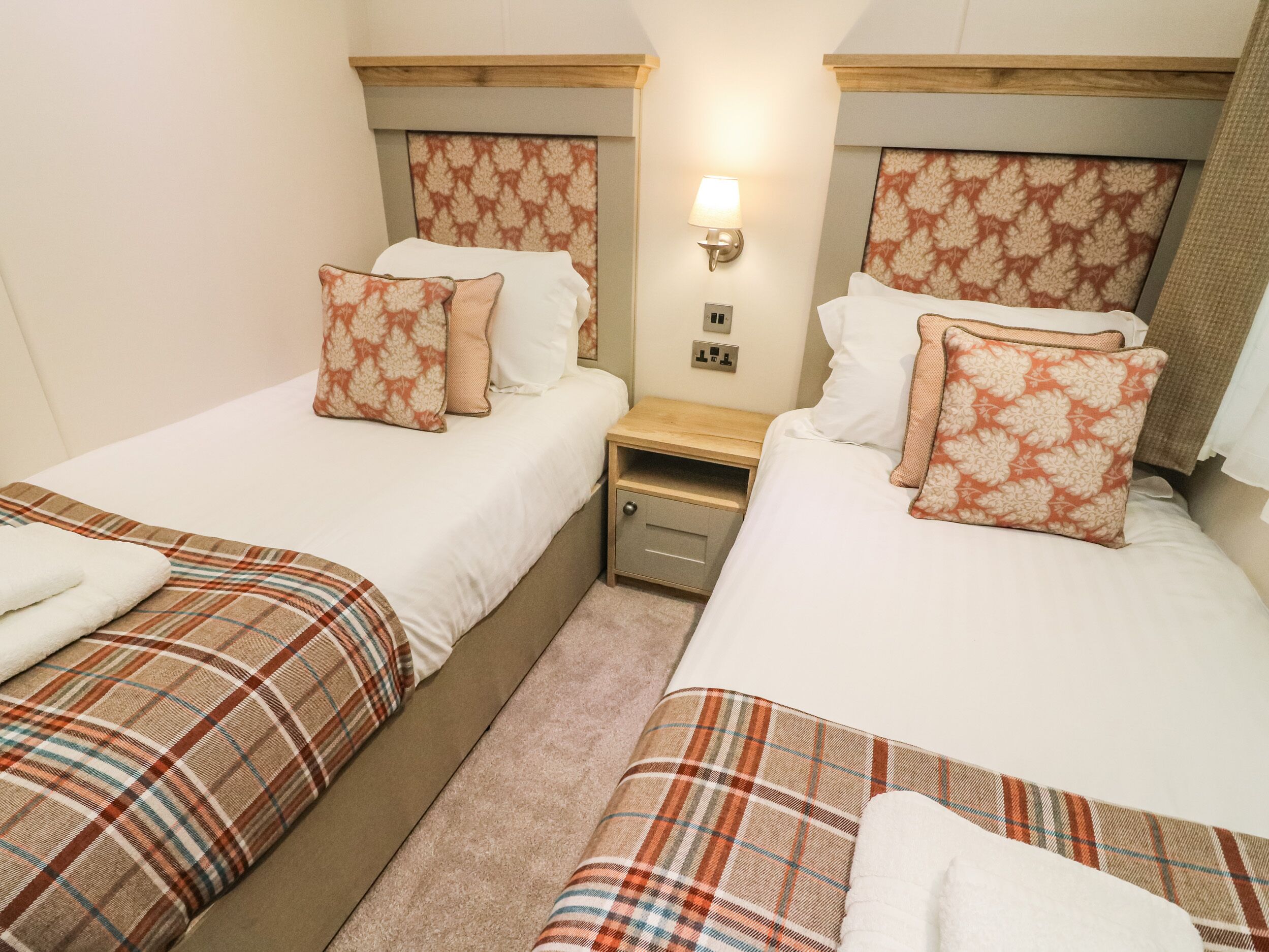 Een hotelkamer met twee bedden, elk met een ruitdessin sprei en bijpassende kussens. Tussen de bedden staat een nachtkastje met een lamp.