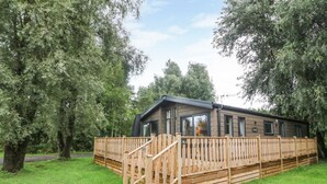 A wooden mobile home with a fenced deck, surrounded by trees and a grassy area.