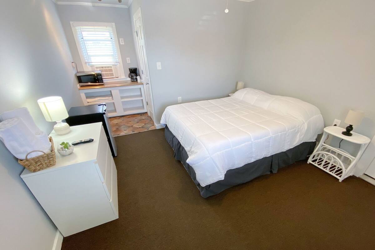 A bedroom with a bed, a desk with a lamp, a small table with a lamp, a basket, and a remote control.