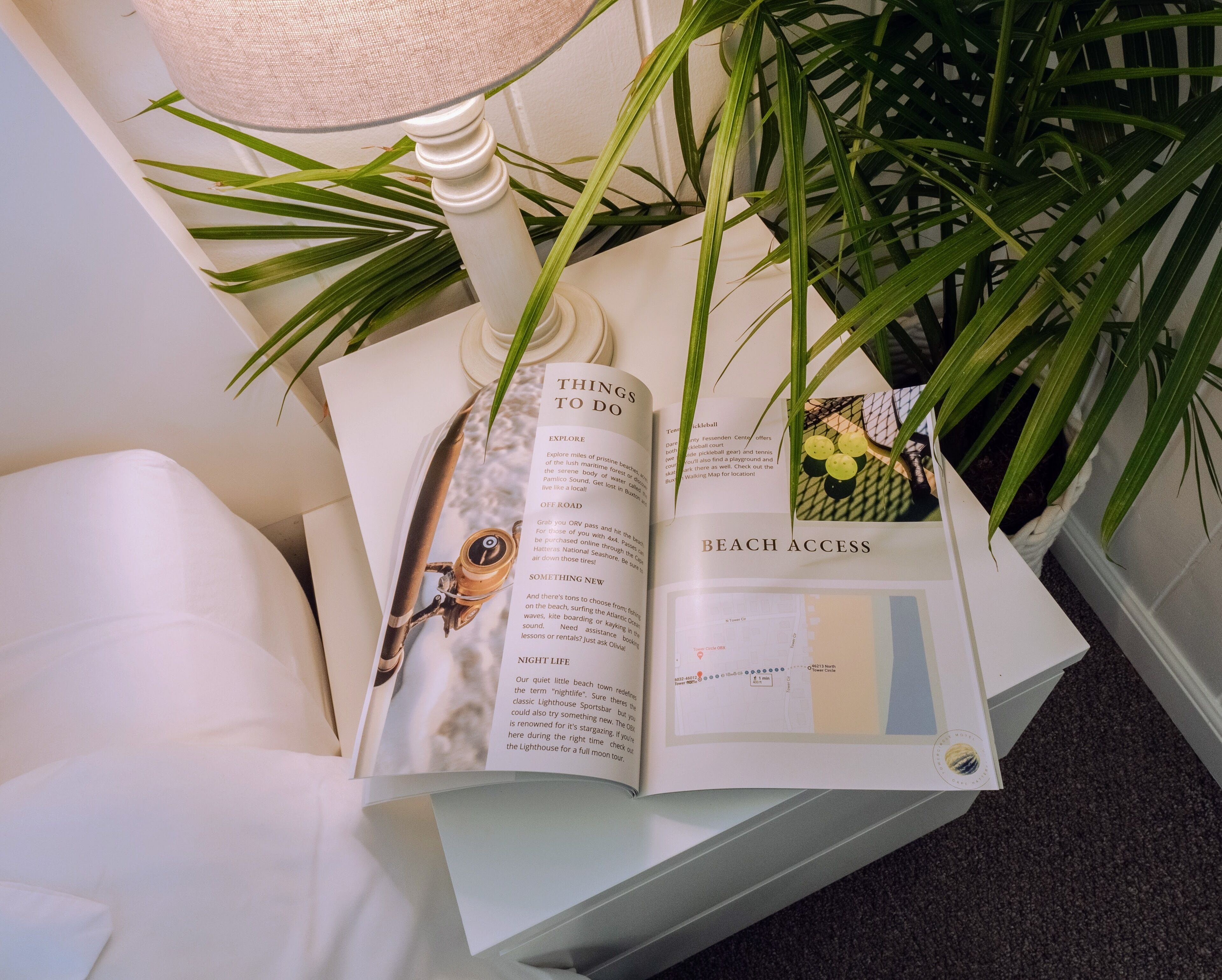 An open brochure with sections titled "Things to do," "Beach Access," and "Night Life," placed on a white table next to a lamp and a potted plant.