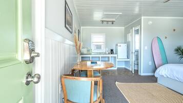 A compact room with a dining area, kitchenette, and a bed with surfboards.