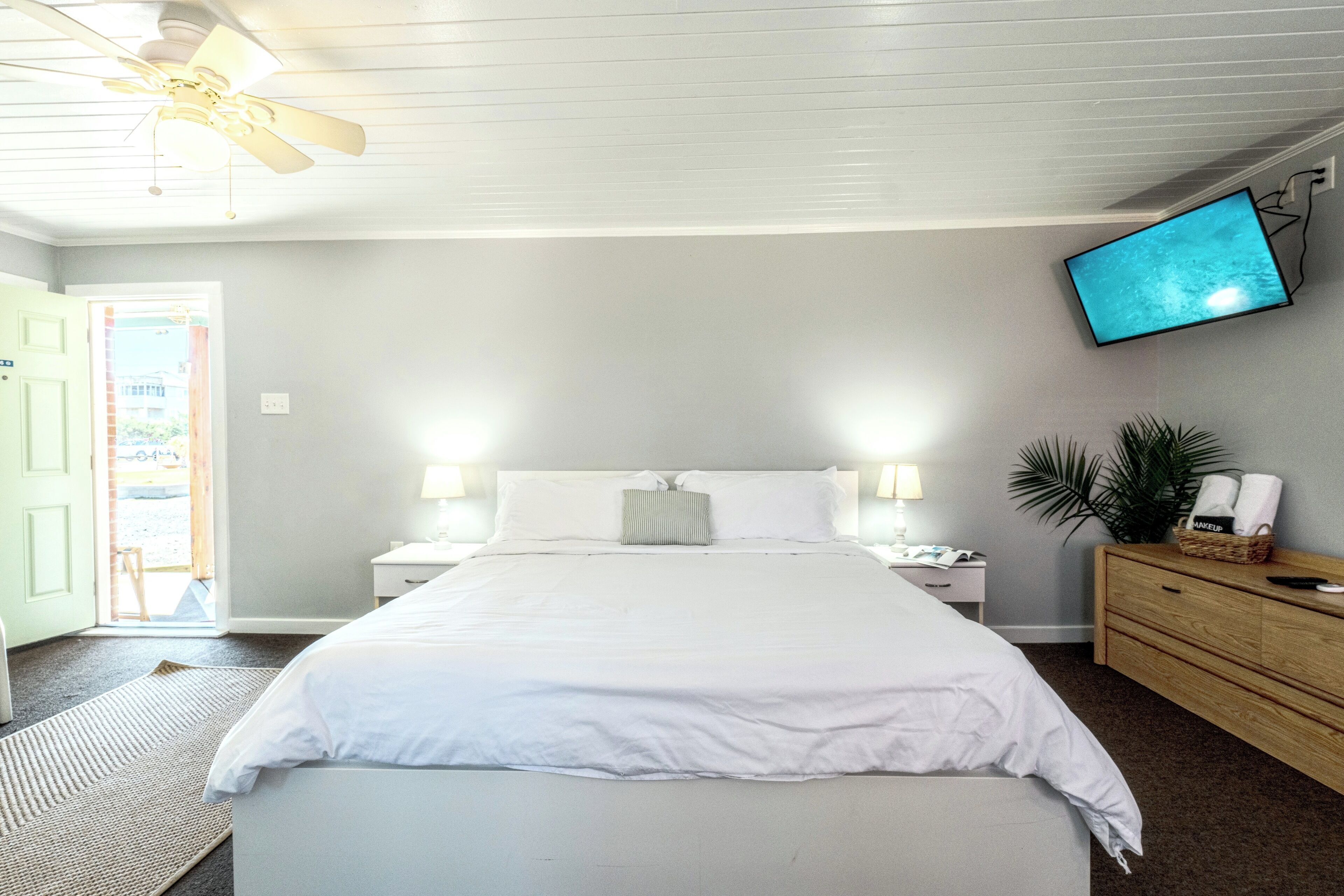 A bedroom with a bed, bedside tables, a TV mounted on the wall, a ceiling fan, and a door leading to an outdoor area.