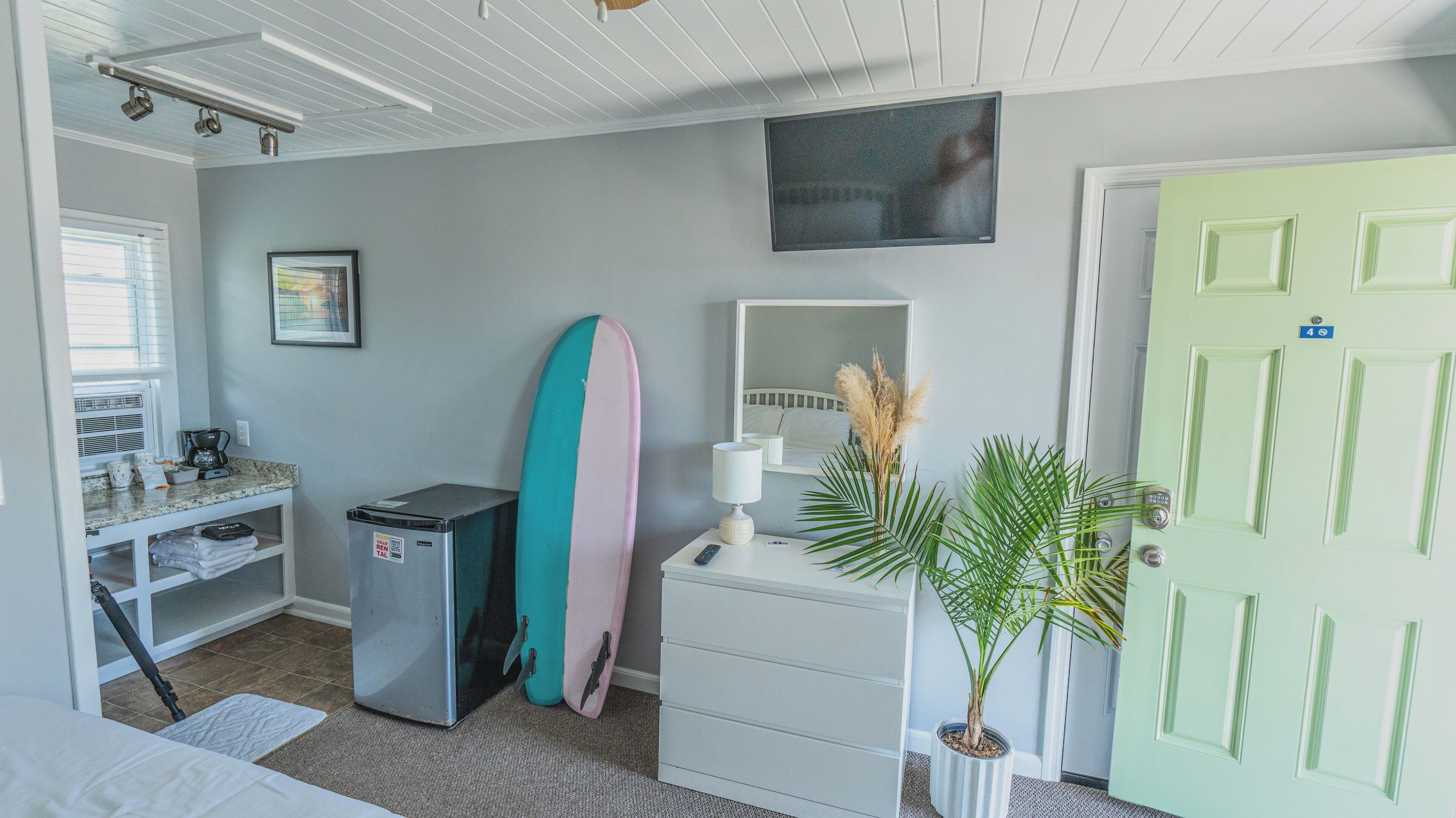 Une chambre à coucher comprenant un lit, une commode, une table de chevet, un bureau avec un ordinateur, une planche de surf, un miroir, une porte et une fenêtre.