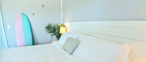A neatly made bed with white linens, a bedside lamp, and a surfboard leaning against the wall.