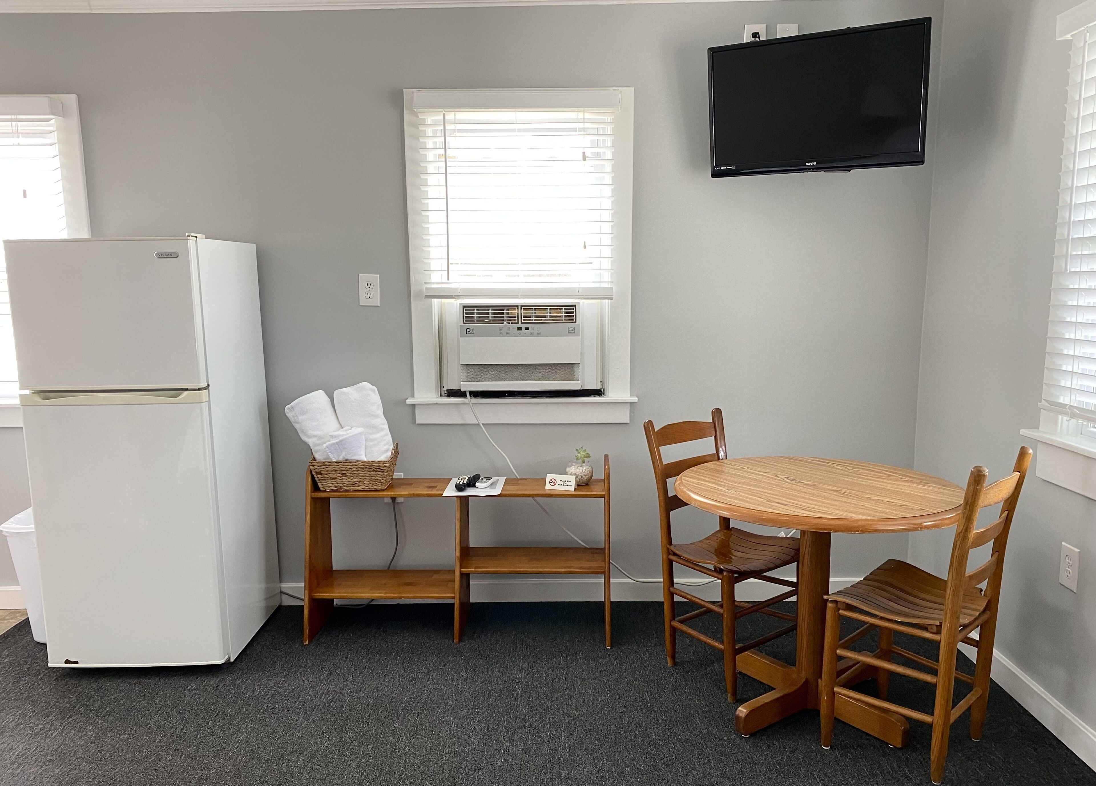 A compact kitchen with a white refrigerator, a wooden dining table with four chairs, a small wicker basket with towels, and a wall-mounted TV.