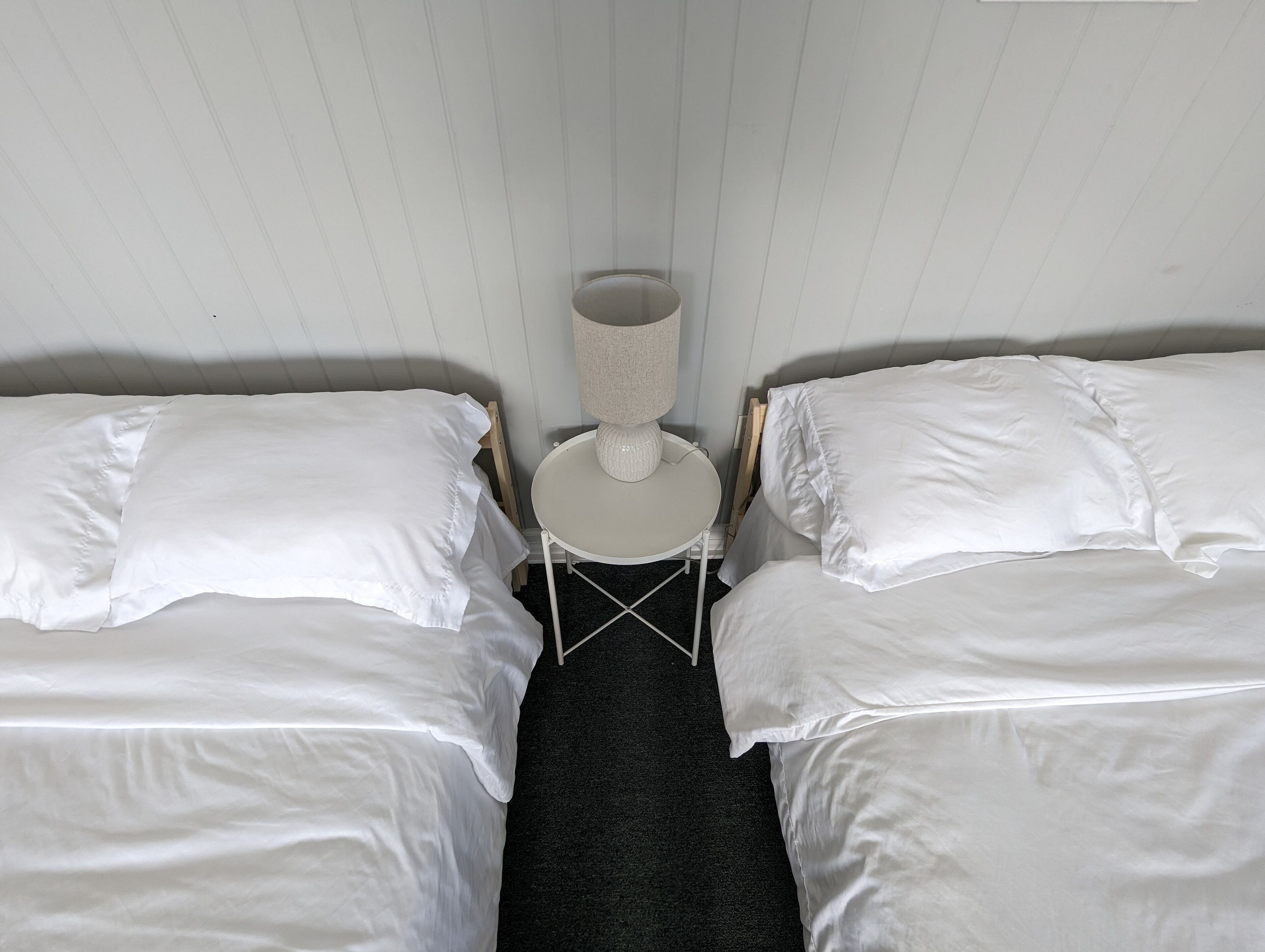 A room with two single beds, each with white bedding, a small round table with a lamp, and a dark carpeted floor.