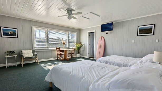 Une chambre d’hôtel avec deux lits, un ventilateur de plafond, une table à manger avec des chaises, une planche de surf et un téléviseur fixé au mur.