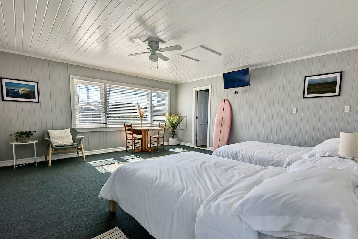 A hotel room with two beds, a ceiling fan, a dining table with chairs, a surfboard, and a TV on the wall.