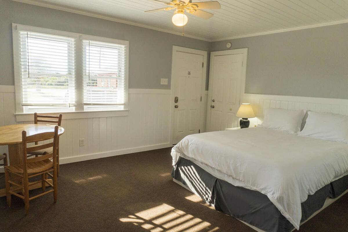 A bedroom with a bed, a desk, and two windows with blinds.
