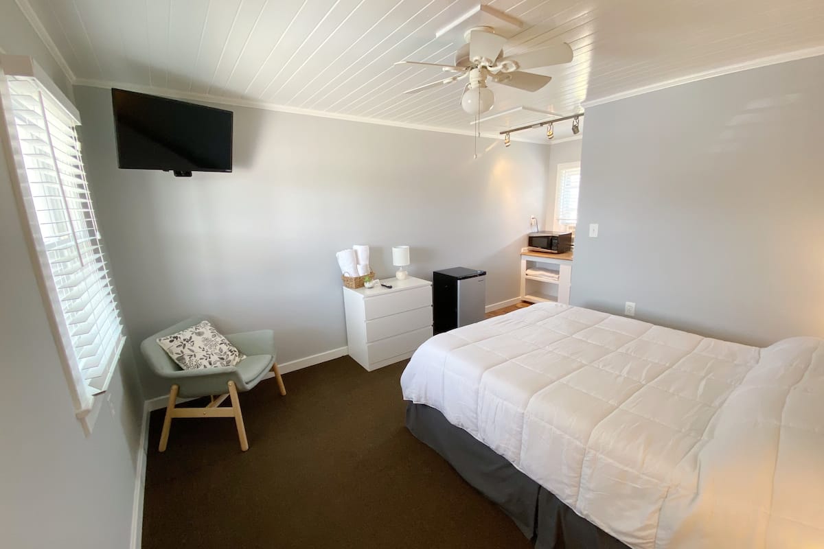 A bedroom with a bed, a desk, a chair, a TV mounted on the wall, and a ceiling fan.