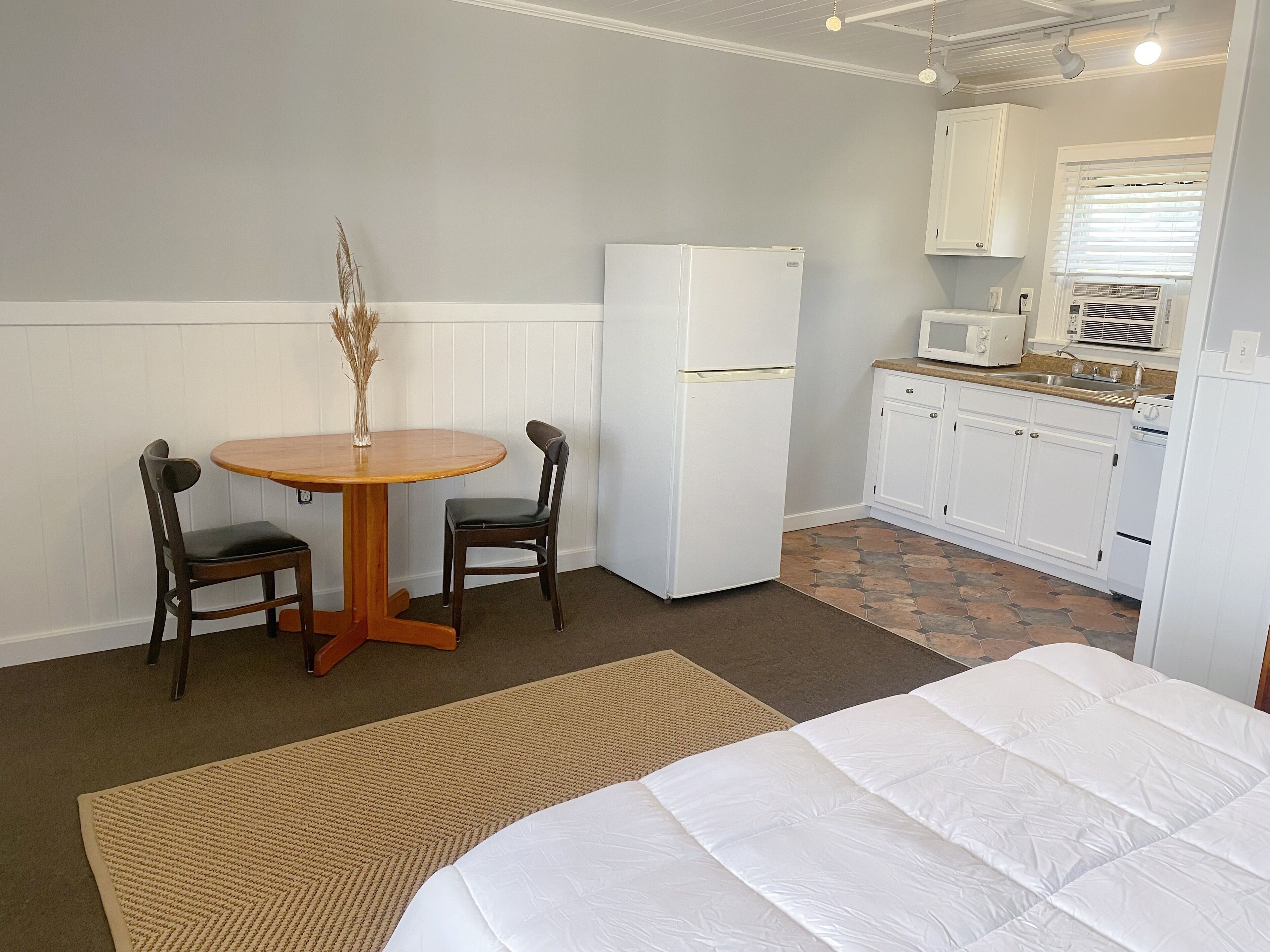Habitación de hotel compacta con cama, una pequeña mesa de comedor con dos sillas, refrigerador, microondas y kitchenette.