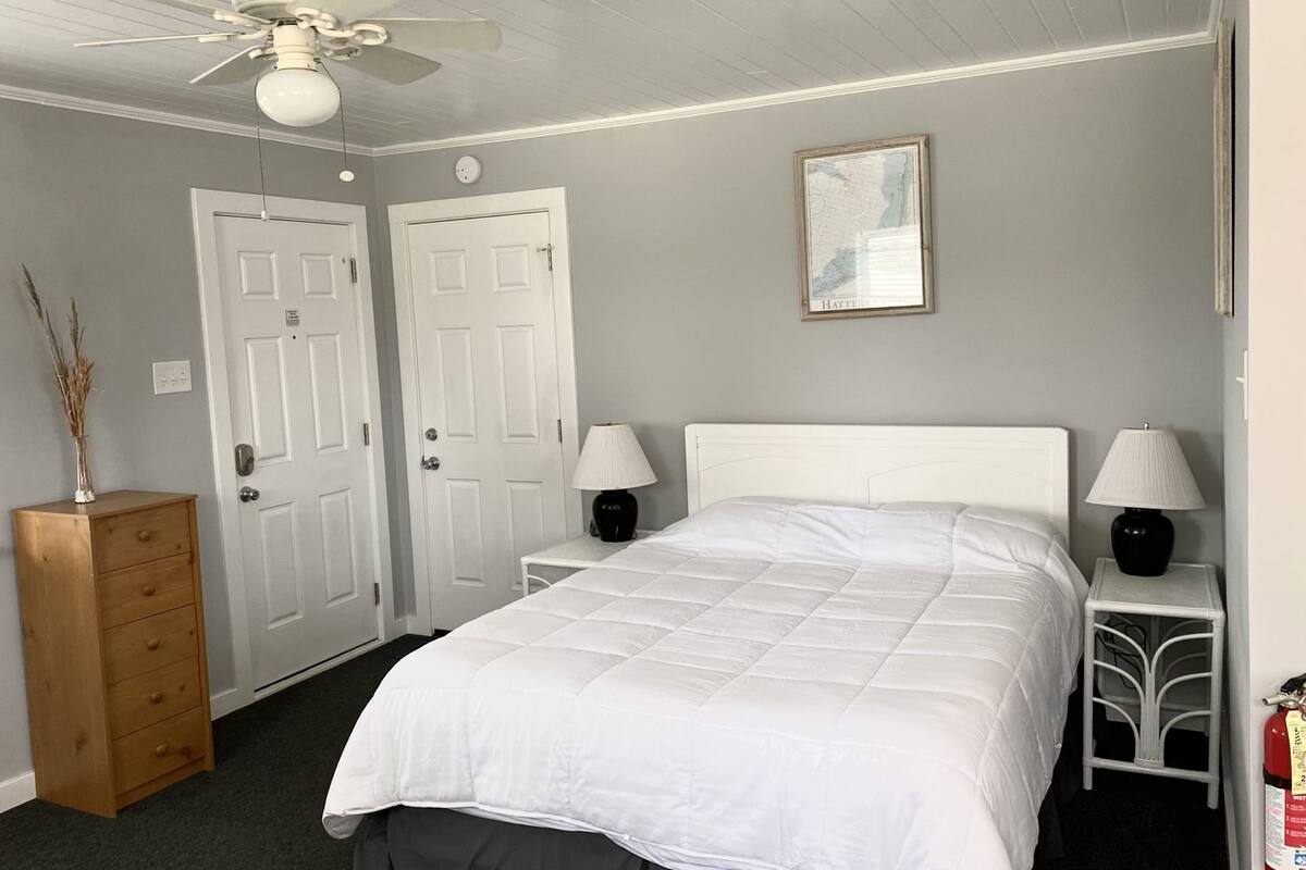 A bedroom with a bed, two bedside tables with lamps, a ceiling fan, two white doors, a wooden dresser, and a framed picture on the wall.