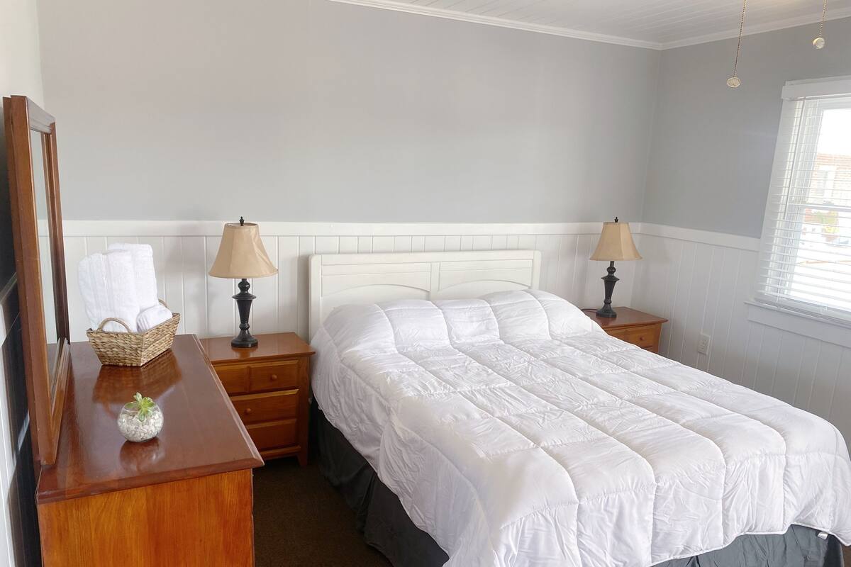 A neatly arranged bedroom with a large bed, two bedside tables, a dresser, and a window with blinds.