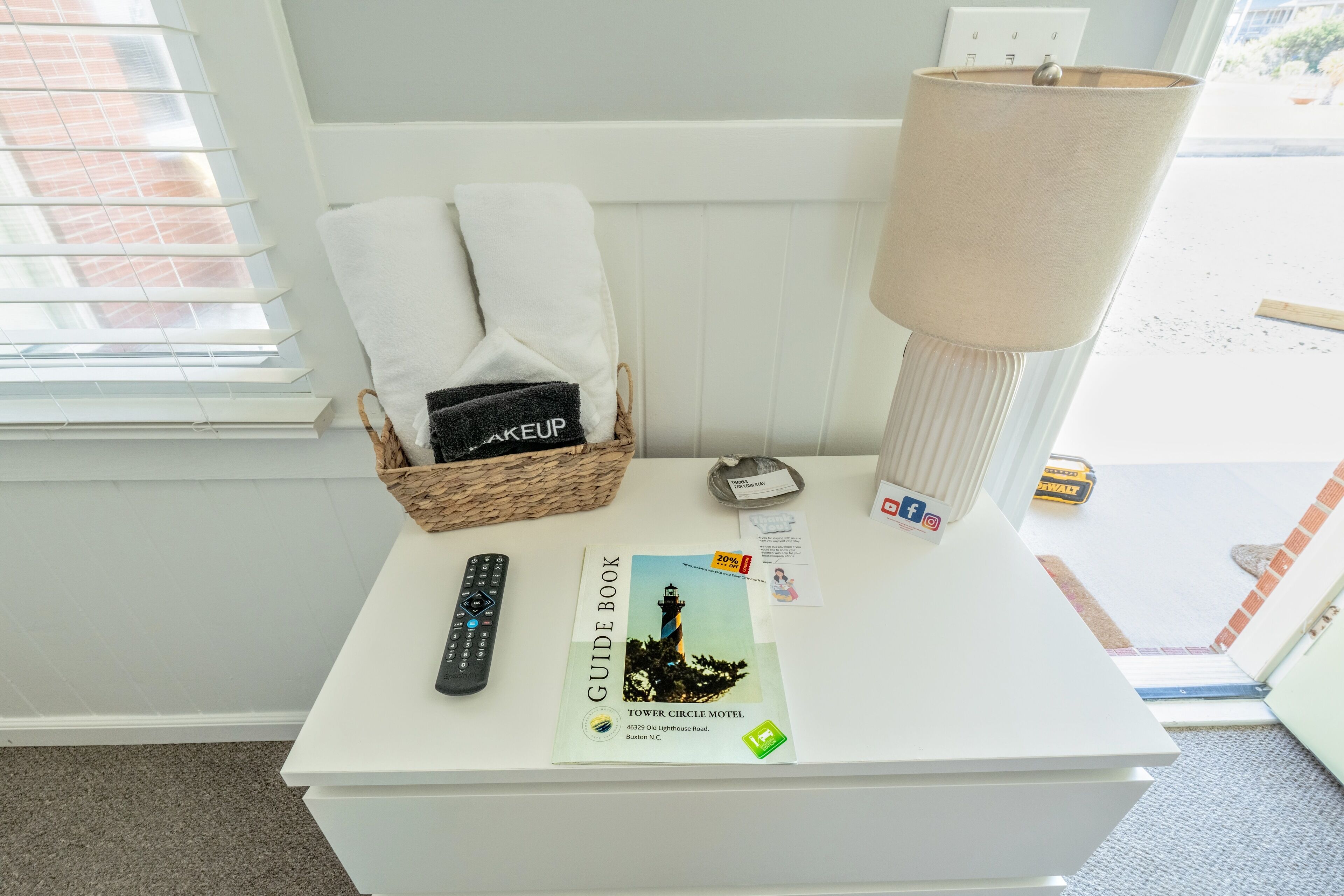 Een witte tafel met een reisgids, een afstandsbediening, een lamp en een mand met handdoeken.